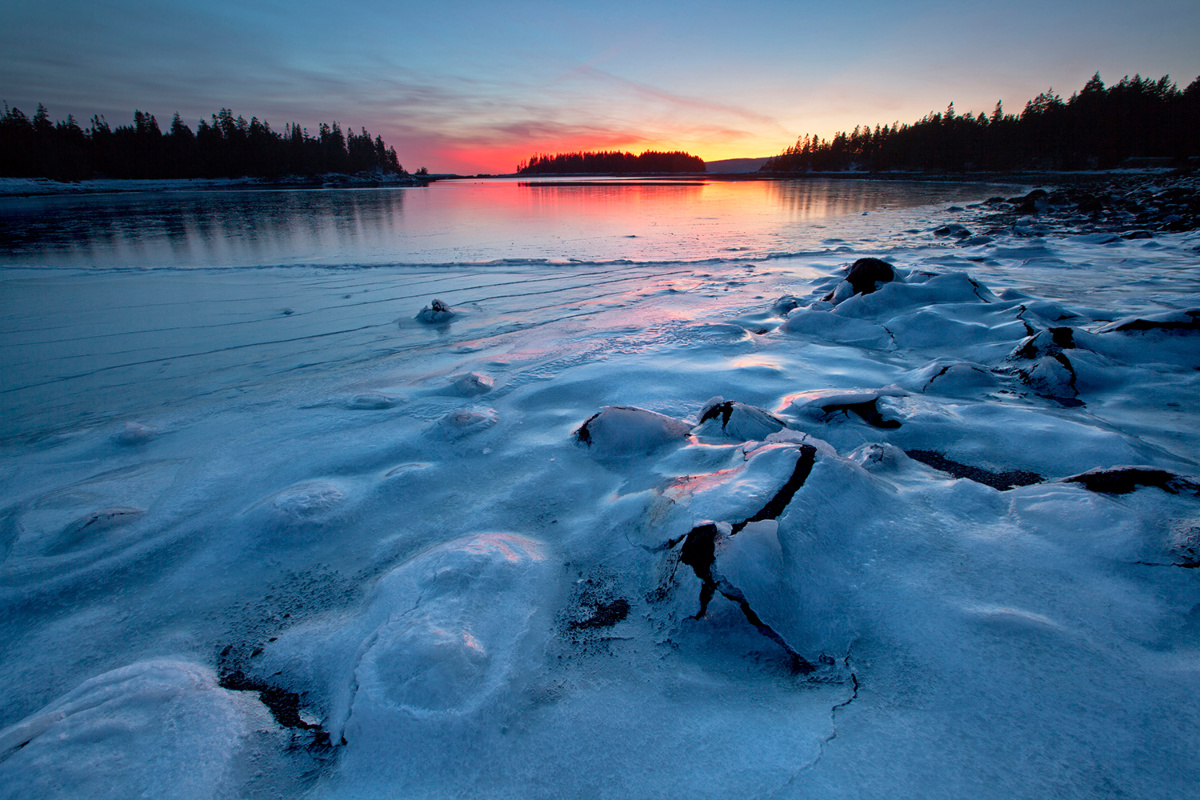 A sheet of ice covers the boulders in West Pond Cove during frigid weather and a fiery sunset on the Schoodic Peninsula, Acadia National Park, Maine, USA